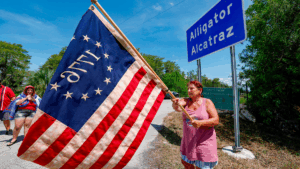 A woman waves a 1776 American flag in front of an Alligator Alcatraz sign in Ochopee, Fla. Photo courtesy of D.A. Varela, The Miami Herald