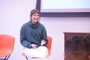 Isabella Gomez Sarmiento of NPR, wearing a green turtleneck sweater and sitting on an orange chair, speaks to fellow journalists.